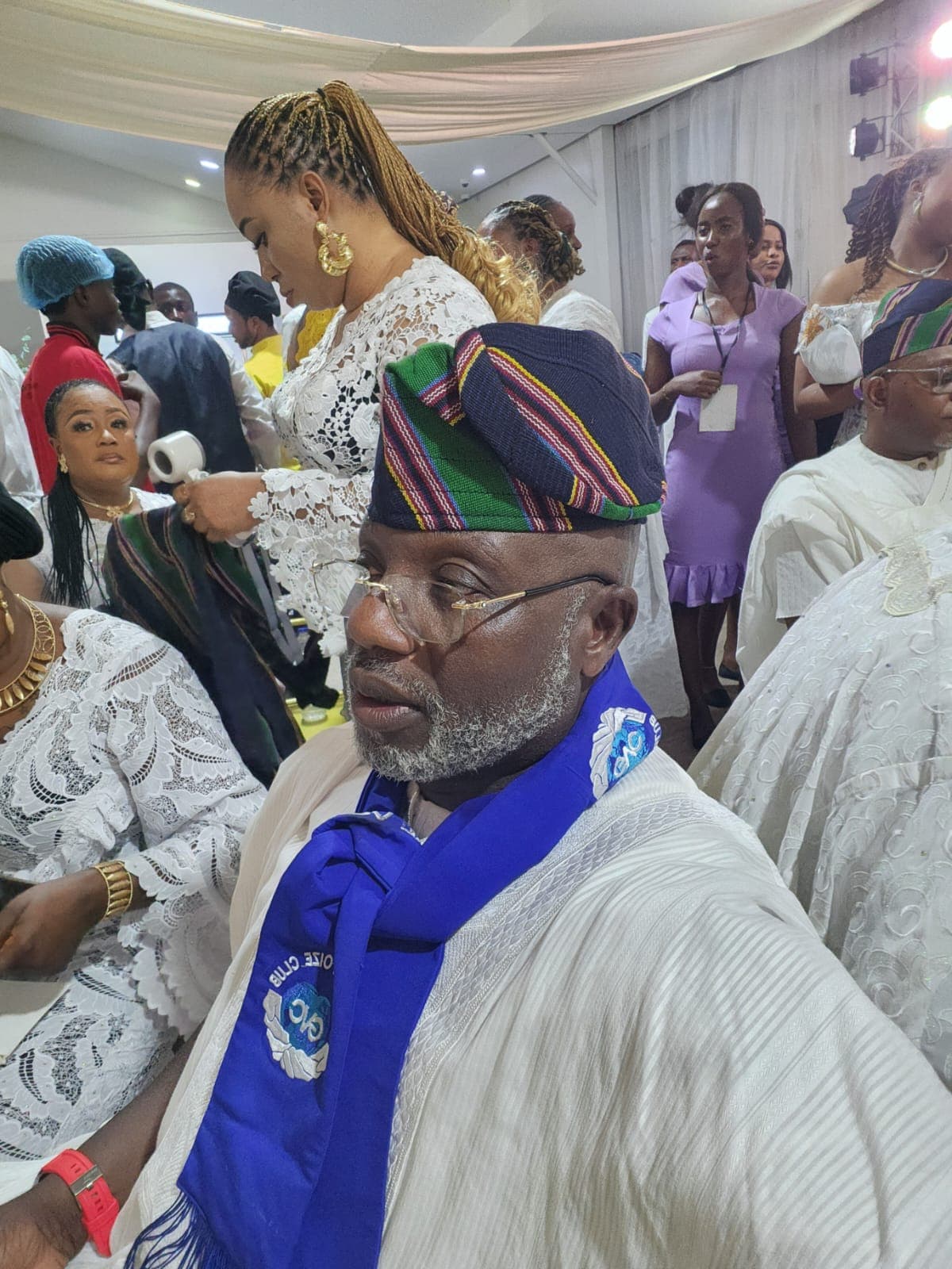 A club member wearing a blue club sash and traditional cap among guests dressed in white lace at a celebration
