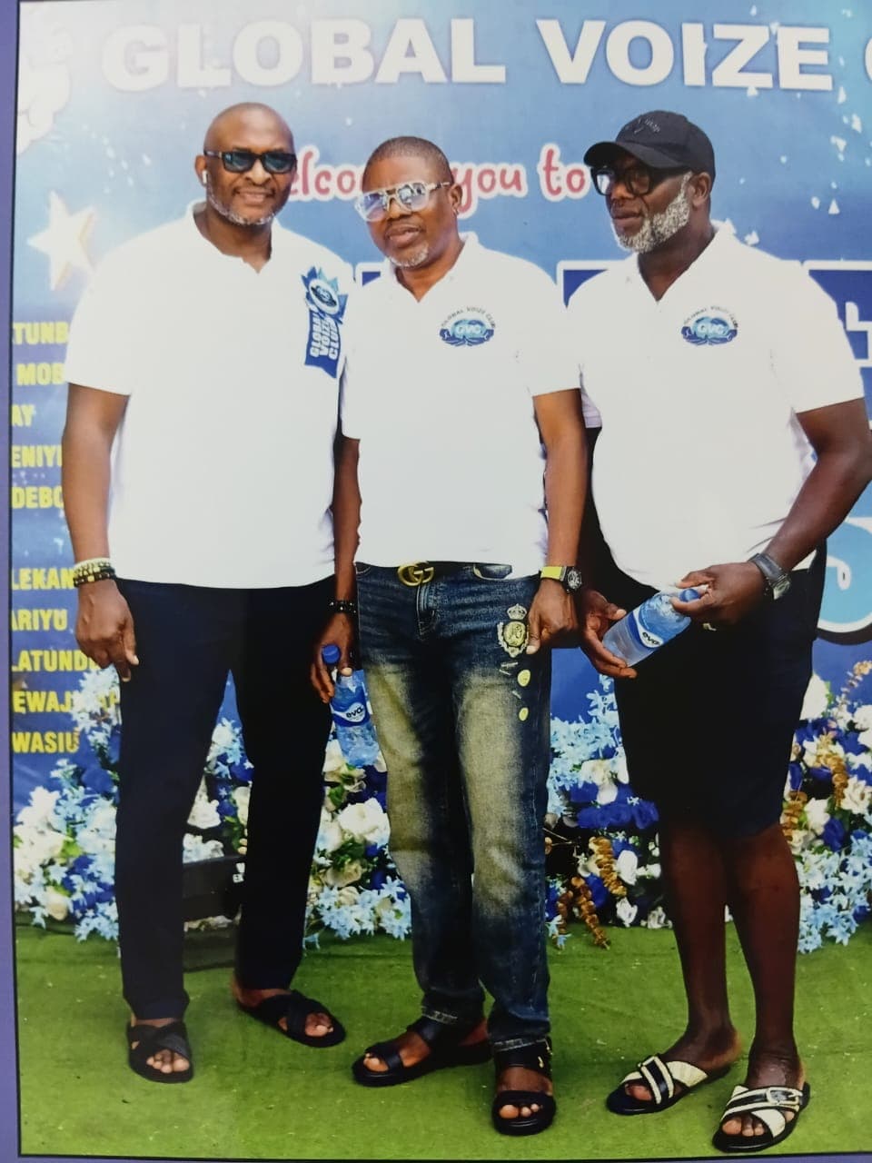 Three club members in white polo shirts holding bottled water in front of the Global Voize welcome banner
